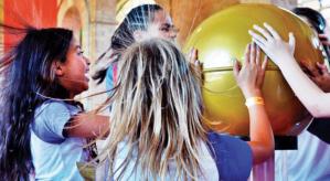 Descrição da imagem: Três meninas com as mãos apoiadas em uma esfera de cobre de cor amarela, chamado Gerador de Van de Graff, que faz seus cabelos se arrepiarem.