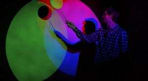 Descrição da imagem: Reflexo de cores verde e rosa na parede em uma sala escura, um homem de perfil segura com a mão direita uma raquete redonda que reflete na parede.