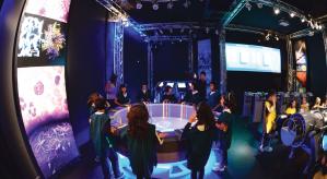 Descrição da imagem: Sala interativa da nanotecnologia.A esquerda, Crianças em pé em um circulo com fones de ouvido jogando em uma tela. A direita, crianças sentadas com fones de ouvido.