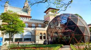Descrição da imagem: Jardim do Museu Catavento. A direita, borboletário que é uma cúpula redonda feita de rede com borboletas voando.