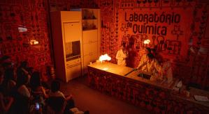 Descrição da imagem: laboratório de química. A esquerda, crianças sentadas de perfil observando uma mulher à direita, de óculos de proteção, jaleco branco e calças preta, as 2 mãos estão esticadas para o alto segurando um bastão em cada mão, e neles está saindo fogo.