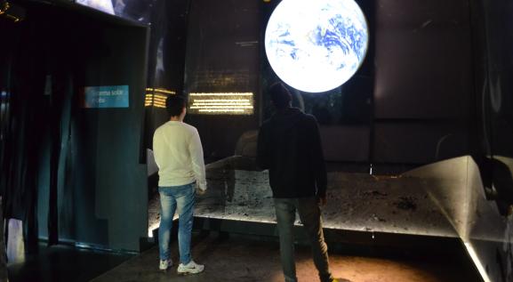 Descrição da imagem: Foto de 2 homens de costas pisando na representação da lua de cor cinza, estão observando a foto do Planeta Terra visto do espaço, a esquerda entrada de uma sala com uma placa azul escrito em letra branca: Sistema Solar e céu