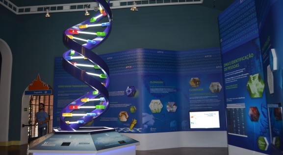 Descrição da imagem: Seção da vida. A esquerda, representação do DNA de dupla hélice em formato helicoidal na vertical acima de uma bancada. A direita, painéis expostos em fundo azul.