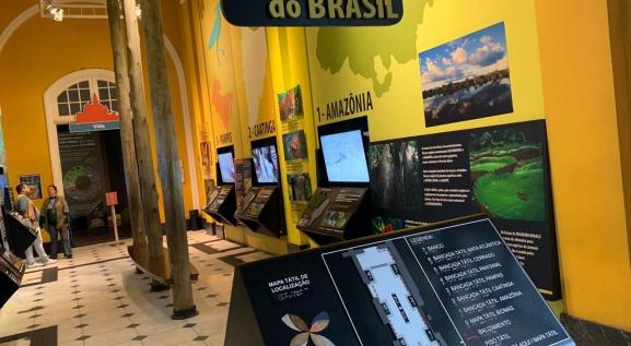 Descrição da imagem: Seção dos Biomas Brasileiros, com painéis e televisões expostas. A direita, escrito: 1 - Amazônia. No centro da imagem uma bancada com mapa tátil de localização.