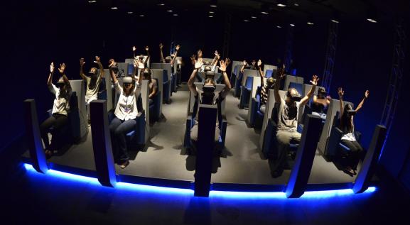 Descrição da imagem: Pessoas sentadas em fileira na vertical, com as duas mãos levantadas usando óculos de realidade virtual. 