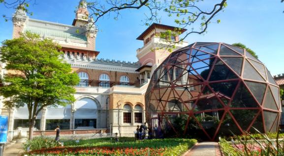 Descrição da imagem: Jardim do Museu Catavento. A direita, borboletário que é uma cúpula redonda feita de rede com borboletas voando.