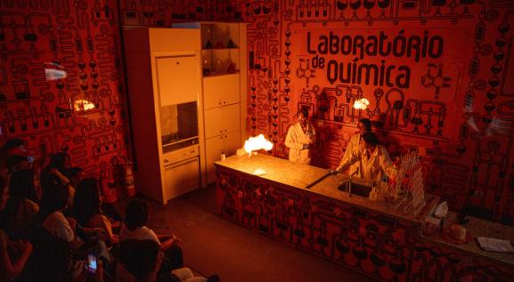 Descrição da imagem: laboratório de química. A esquerda, crianças sentadas de perfil observando uma mulher à direita, de óculos de proteção, jaleco branco e calças preta, as 2 mãos estão esticadas para o alto segurando um bastão em cada mão, e neles está saindo fogo.