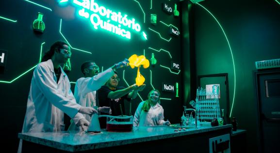 Descrição da imagem: laboratório de química. quatro pessoas com jaleco branco olhando para uma que está com a mão estendida e tem fogo saindo dela, devido a um experimento químico