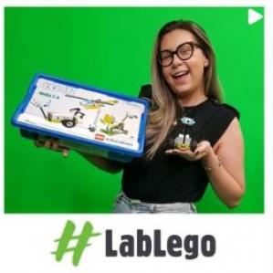 Fundo verde e a frente uma mulher loira, segurando uma caixa de peças de Lego azul