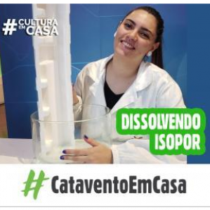 Foto de uma mulher com jaleco branco sorrindo segurando um isopor. Ao fundo uma parede na cor azulada