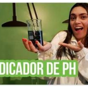 Foto tirada com cenário de laboratório, uma moça segura um frasco na mão enquanto indica esse frasco sorrindo. Na parte inferior da foto escrito: "Indicador de PH"